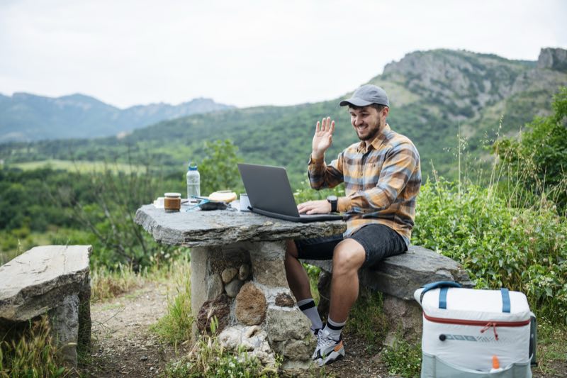 Person arbeitet mit Laptop im Freien und winkt in eine Videokonferenz