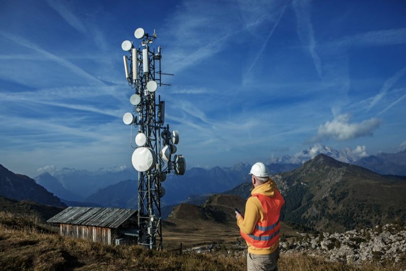 Techniker mit Schutzhelm vor einem Mobilfunkmast in den Bergen